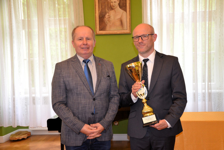 Dwóch mężczyzn w garniturach. Jeden z nich trzyma złoty puchar.