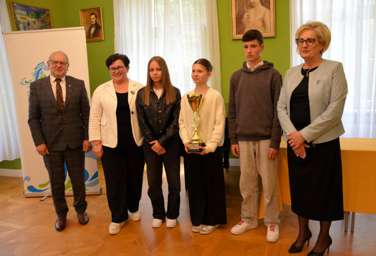 Grupa ludzi stoi w sali. Kobieta w środku grupy trzyma złoty puchar.