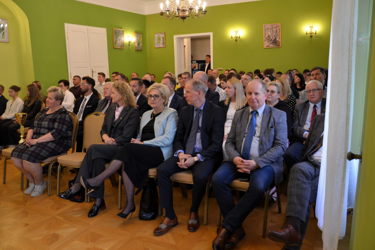 Widownia siedzi w sali z zielonymi ścianami.