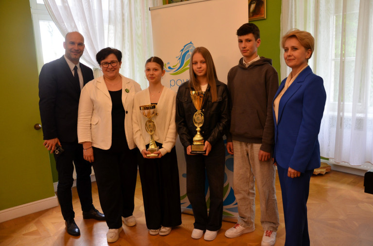 Grupa ludzi stoi w sali. Dwie kobiety trzymają puchary.