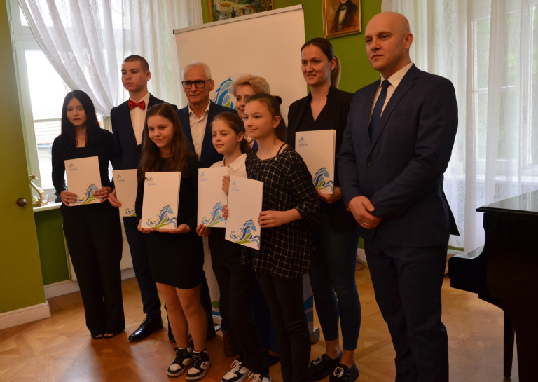 Grupa ludzi stoi na tle fortepianu. Część z nich trzyma dokumenty.