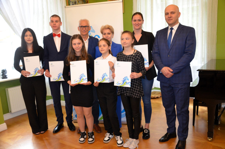 Grupa ludzi stoi na tle fortepianu. Część z nich trzyma dokumenty.